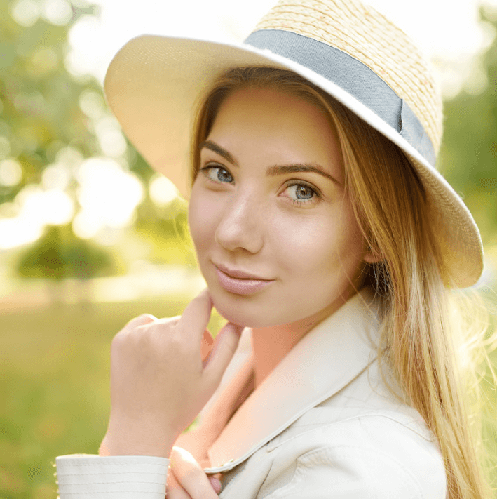 Mujer luciendo un sombrero, sonriendo suavemente, en un entorno natural con luz cálida, reflejando confianza y belleza.